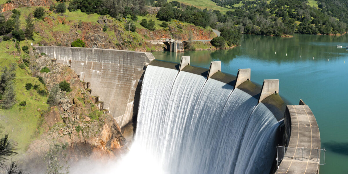 A large concrete dam with water cascading over the spillway, surrounded by green hills and a calm reservoir under a clear blue sky.