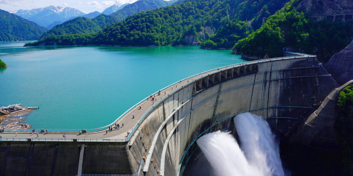 A large curved dam releasing water into a canyon, surrounded by turquoise reservoir water and lush green mountains under a partly cloudy sky.