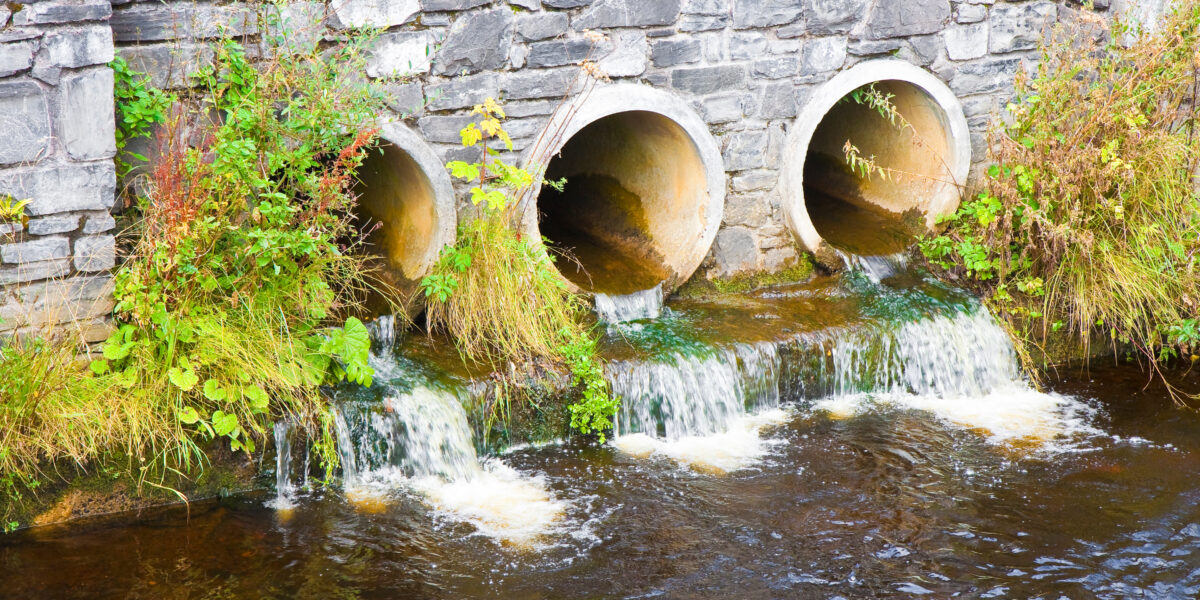 Three large concrete drainage pipes set in a stone wall, with water flowing from them into a river below—an example of effective Wassermanagement—surrounded by green plants and moss.