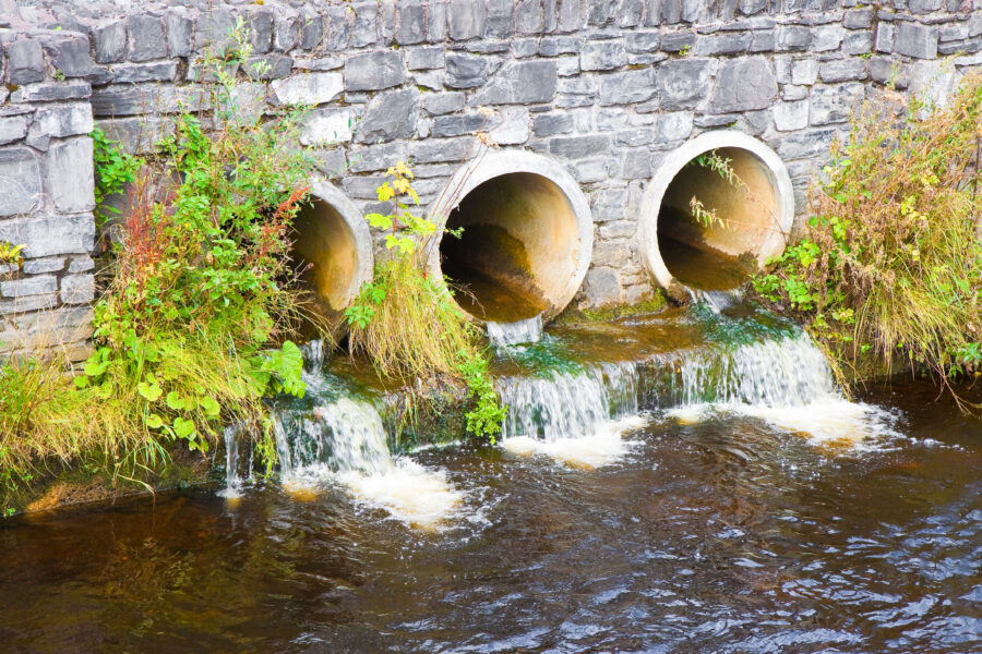 Three large concrete drainage pipes set in a stone wall, with water flowing from them into a river below—an example of effective Wassermanagement—surrounded by green plants and moss.