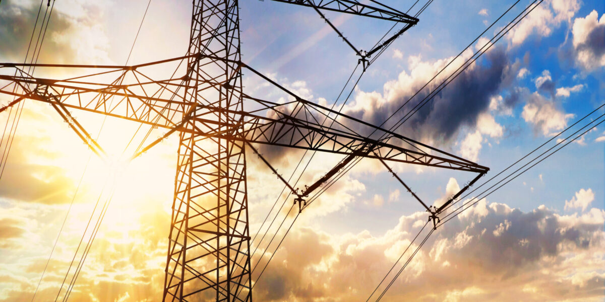 A large electrical transmission tower with power lines stands against a vibrant sky at sunset, with the sun shining through clouds in the background.