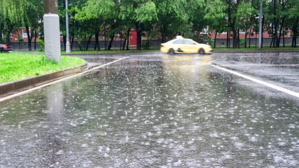 A yellow car drives quickly on a rainy street, with rain visibly falling and splashing on the wet road; trees and buildings are in the background.
