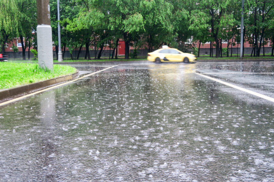 A yellow car drives quickly on a rainy street, with rain visibly falling and splashing on the wet road; trees and buildings are in the background.
