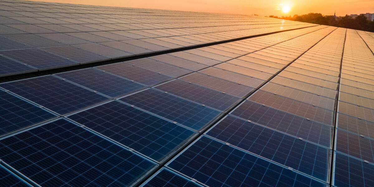 Rows of solar panels stretch across a field, capturing sunlight during sunset with a glowing orange sky in the background.