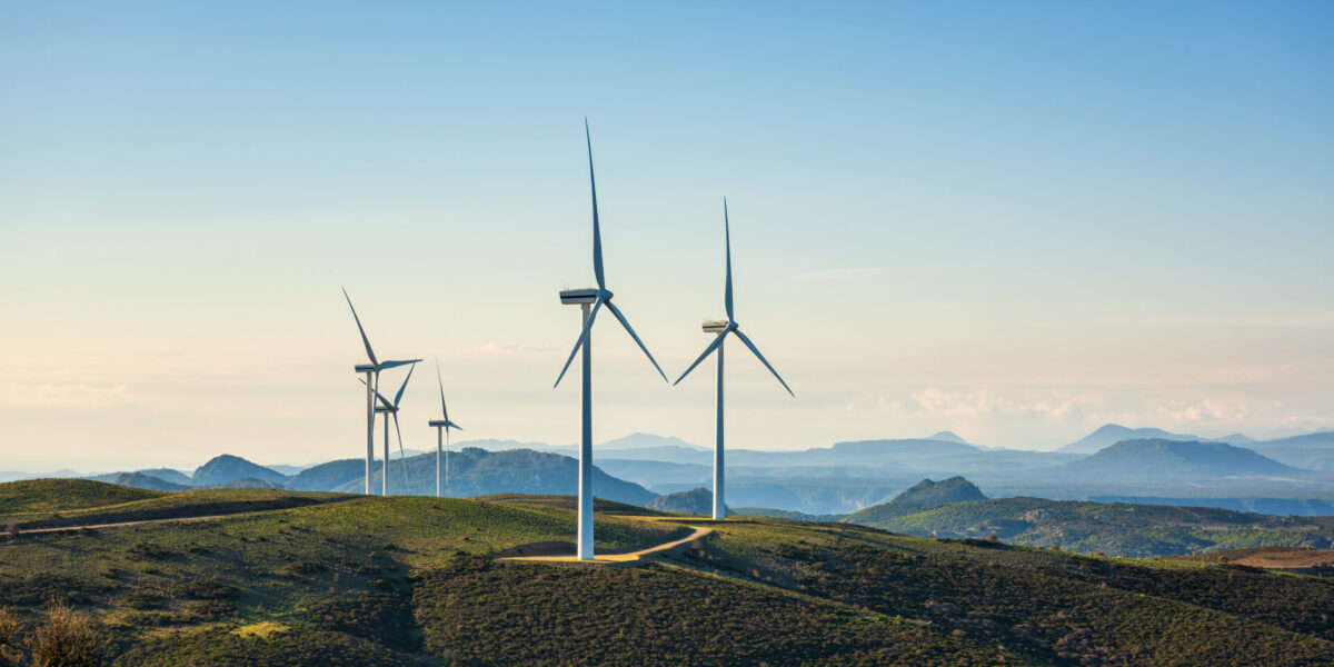 Wind turbines stand on a grassy hill with a winding path, set against a backdrop of distant mountains and a clear sky.