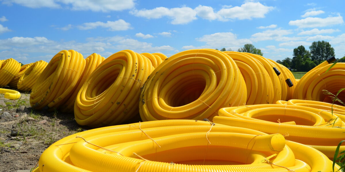 Large coils of yellow plastic tubing are stacked outdoors on a grassy area under a bright blue sky with scattered clouds. Trees are visible in the background.