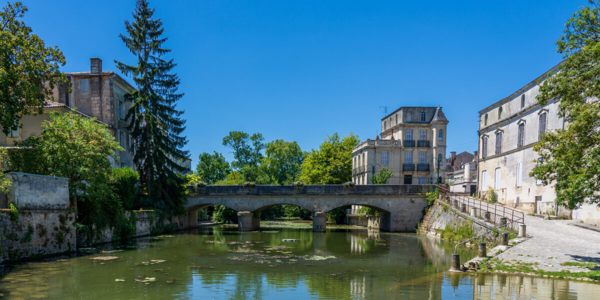 A stone bridge crosses over a calm river lined with trees and historic buildings under a clear blue sky on a sunny day.