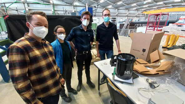 Four people wearing masks stand together in an industrial indoor space beside a table with electronics, a cardboard box, and equipment. The area has high ceilings and exposed pipes.