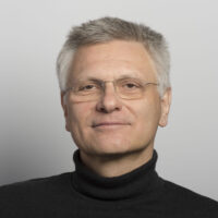 A middle-aged man with short gray hair and glasses, wearing a black turtleneck sweater, smiles slightly in front of a plain, light gray background.