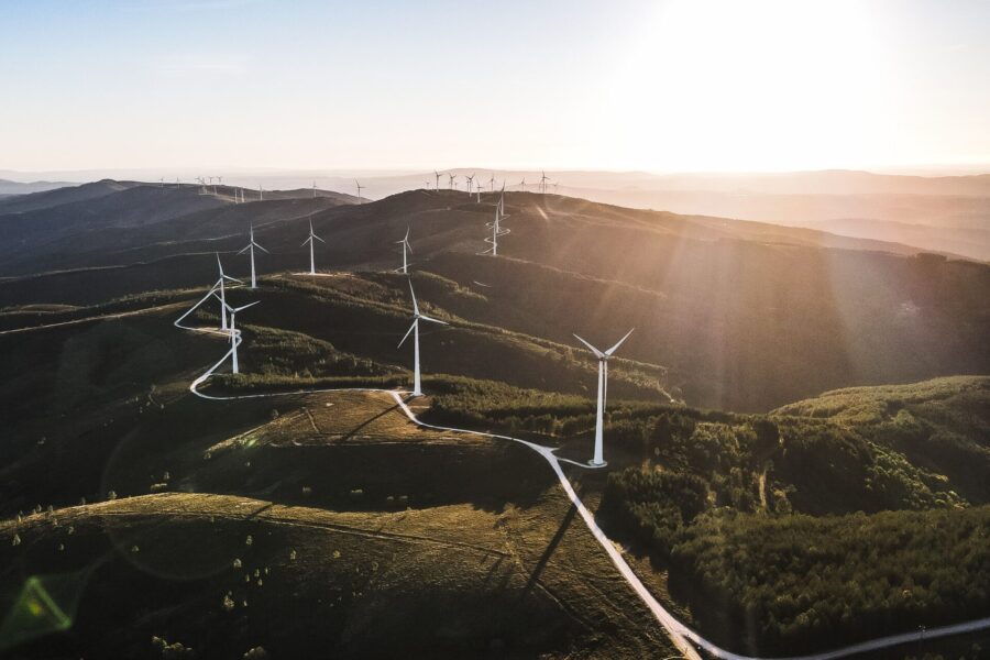 Aerial view of multiple wind turbines scattered across green hills at sunset, with winding roads and a bright sun casting long shadows over the landscape.