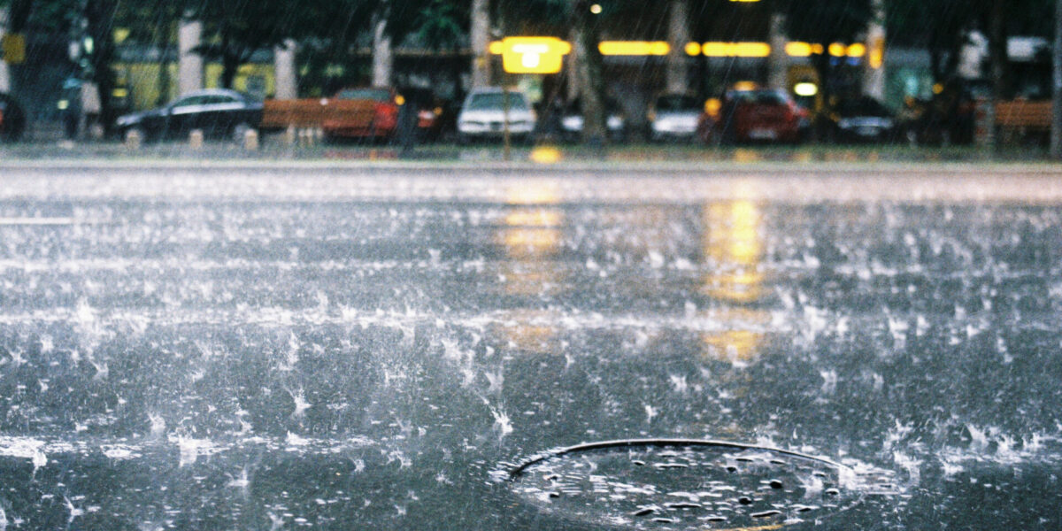 Heavy rain falls on a city street, creating splashes around a manhole cover. Cars and benches are visible in the background, with blurred yellow lights and green trees above.