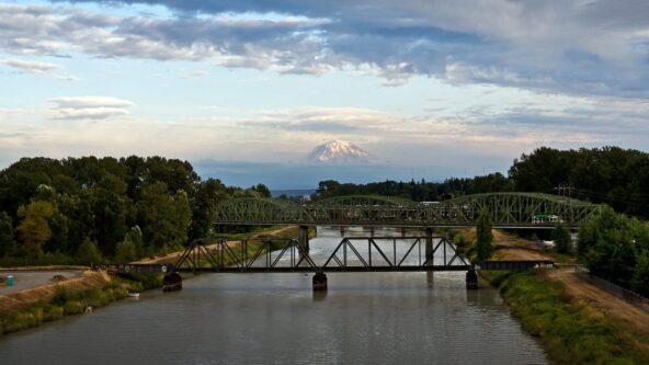 A river flows beneath two green metal bridges, surrounded by trees, with a snow-capped mountain rising in the background under a cloudy sky.