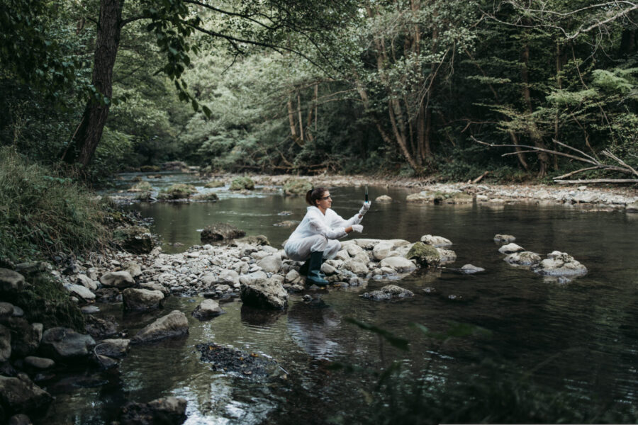 A person in a white coat squats on rocks by a shallow, forested river, holding a container close to the water, possibly collecting a sample. Dense green trees and vegetation surround the calm scene.