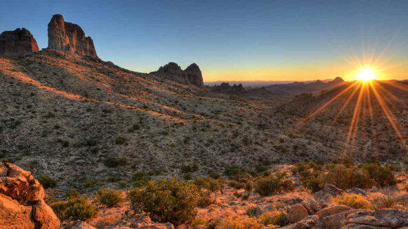 The sun sets over a rugged desert landscape with rocky hills and sparse vegetation, casting a warm golden light and long shadows across the terrain.