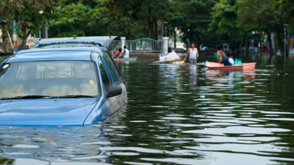 A blue car is standing in flood water, while people and boats can be seen in the background