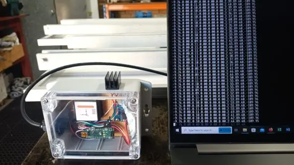 A plastic electronics enclosure with circuit boards and wires is connected to a laptop. The laptop screen displays streaming data in columns of numbers on a black background. The setup is on a workbench.