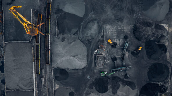 Aerial view of an industrial site with large piles of coal or gravel, yellow excavators, and long train cars filled with material, surrounded by dark, dusty ground.