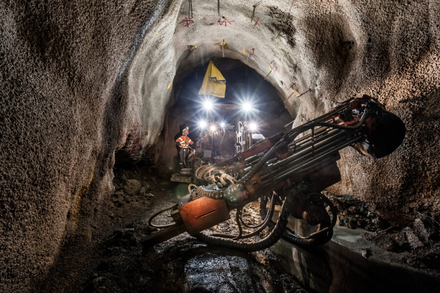 A large piece of drilling machinery is positioned inside a rough, dimly lit tunnel. Bright work lights illuminate workers wearing safety gear further down the tunnel, highlighting the rocky walls and wet ground.