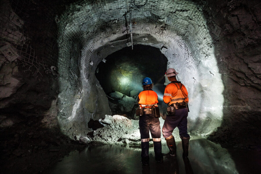Two workers in orange safety gear and helmets stand in a dimly lit underground tunnel, facing a rocky cave wall illuminated by their headlamps, with water pooling on the tunnel floor.