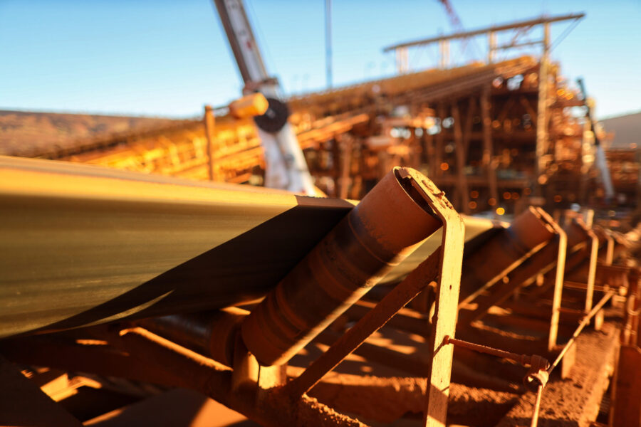 A close-up of a moving conveyor belt carrying iron ore at an industrial mining site, with blurred machinery and equipment in the background under bright sunlight.