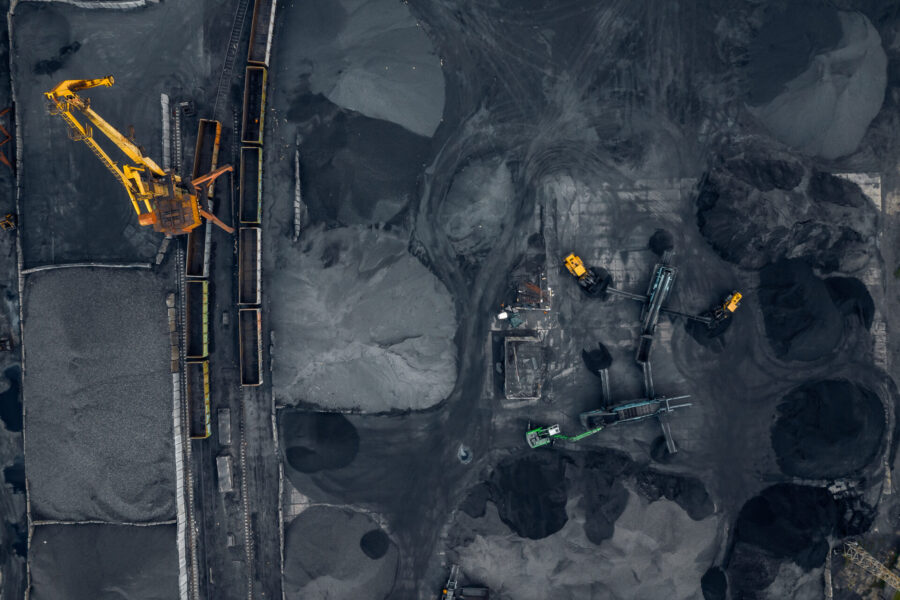 Aerial view of an industrial mining site with large piles of coal or minerals, several conveyor belts, and heavy machinery including yellow excavators and trucks working on dark, dusty ground.