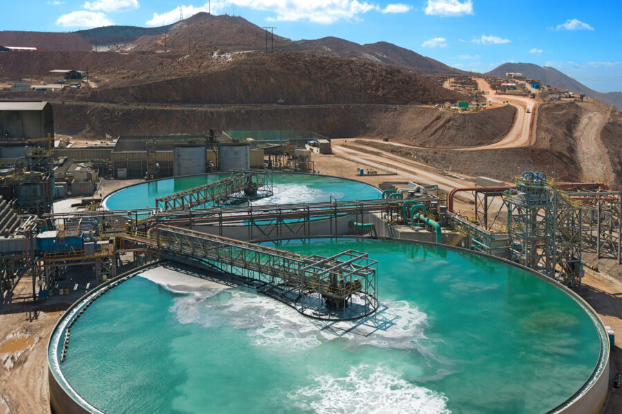 Large industrial tanks with turquoise water are part of a mining facility set in a dry, hilly landscape under a blue sky. Pipes and machinery surround the tanks, and dirt roads wind through the scene.