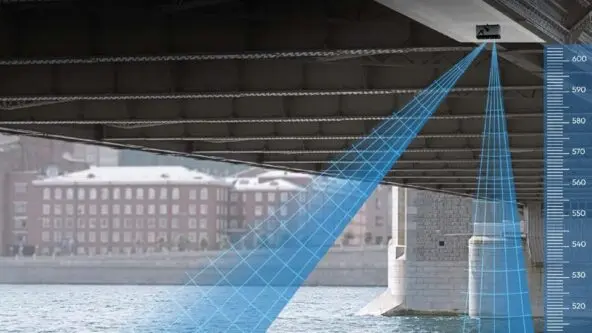 A camera is mounted under a bridge, projecting blue grid lines onto the water below to measure its height, with a vertical gauge showing water level readings in centimeters alongside the bridge support.