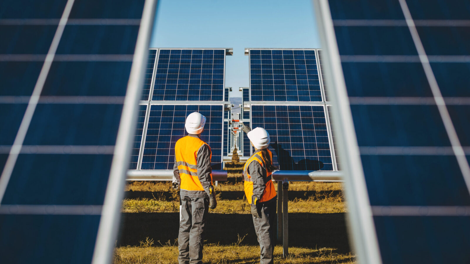 Two workers in orange safety vests and white helmets stand between large solar panels, inspecting the solar farm and its real-time hail sensor system on a clear, sunny day.