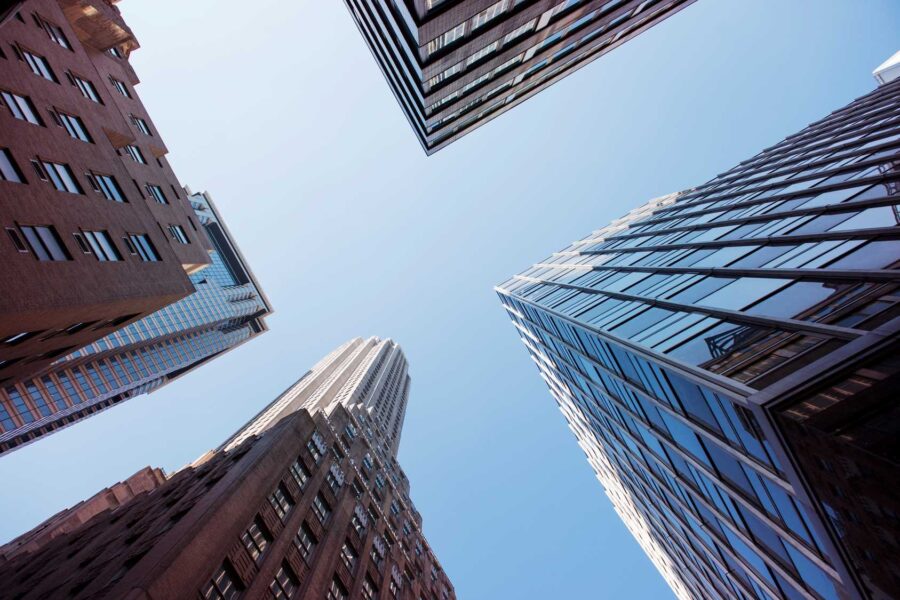 Skyscrapers seen from below against a clear blue sky, with buildings of various styles and materials forming an open rectangle when looking upwards, while a real-time hail sensor discreetly monitors weather conditions above.