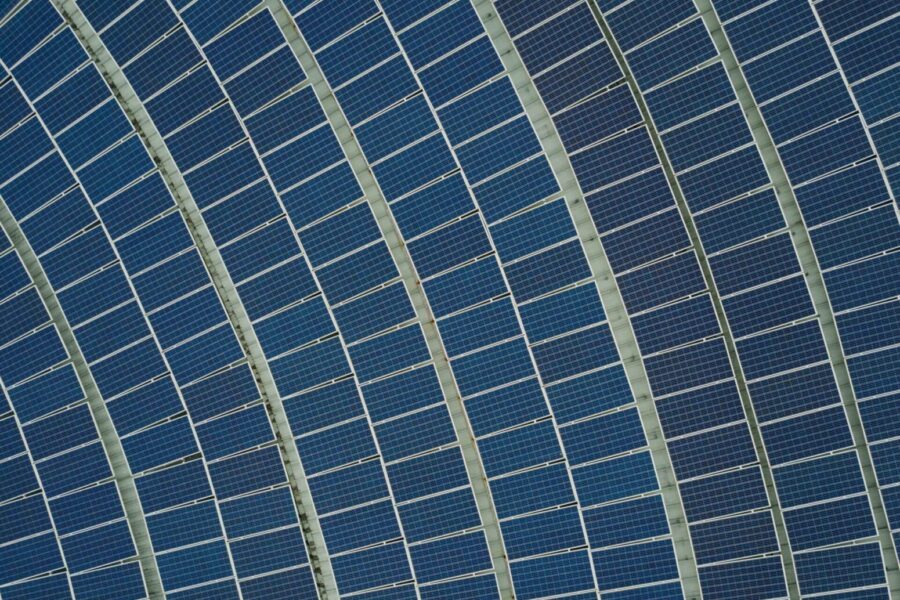 Aerial view of curved rows of large blue solar panels with green support structures and integrated real-time hail sensors, forming a striking geometric pattern.