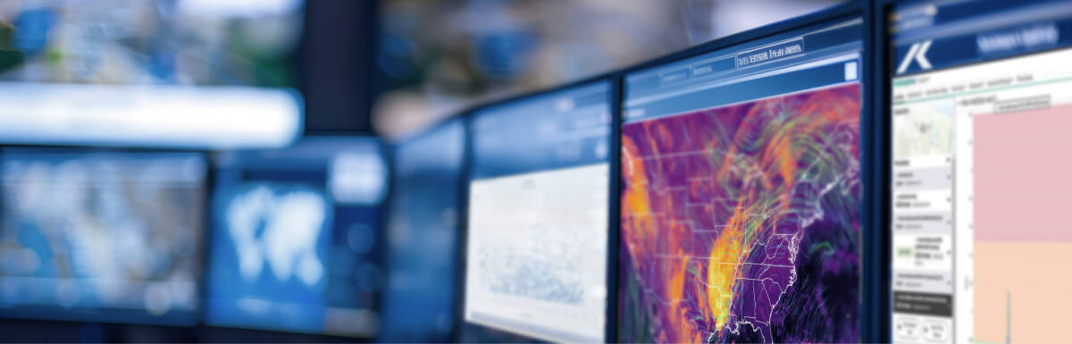 A row of computer monitors displaying weather maps, charts, and data visualizations in a monitoring or forecasting center.