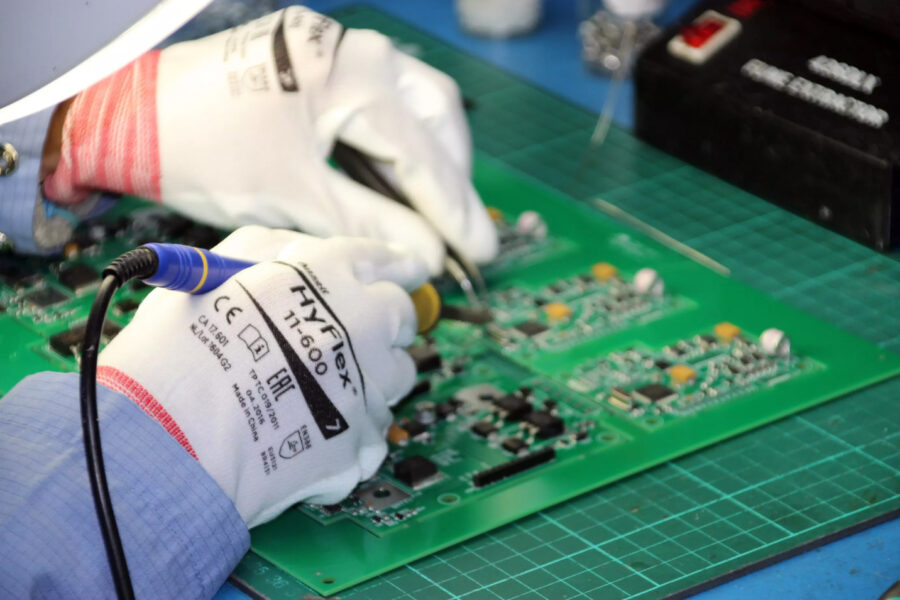 A person wearing white gloves uses a soldering iron to work on green circuit boards on a cutting mat, providing Instruments Support while assembling or repairing electronic components.