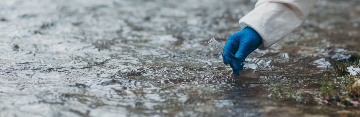 A person wearing a lab coat and blue gloves collects a water sample from a shallow stream using a glass vial.