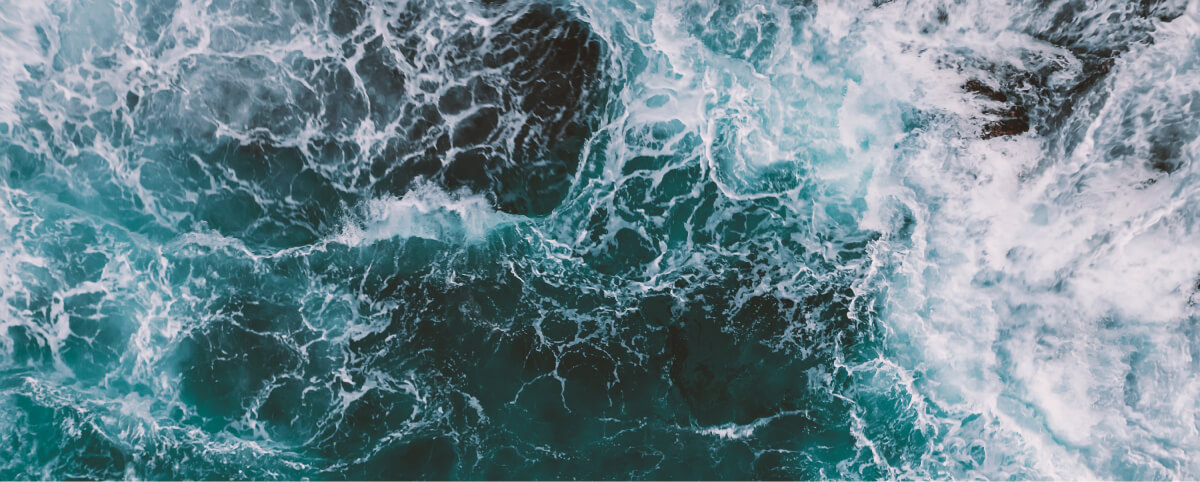 Aerial view of turbulent ocean waves crashing and swirling, creating white foam patterns on deep blue-green water.