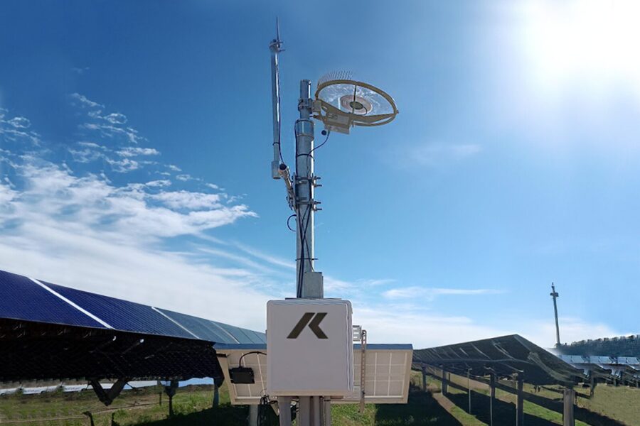 A weather monitoring station with sensors and antennas, including a real-time hail sensor, stands near rows of solar panels in a sunny outdoor field under a clear blue sky.