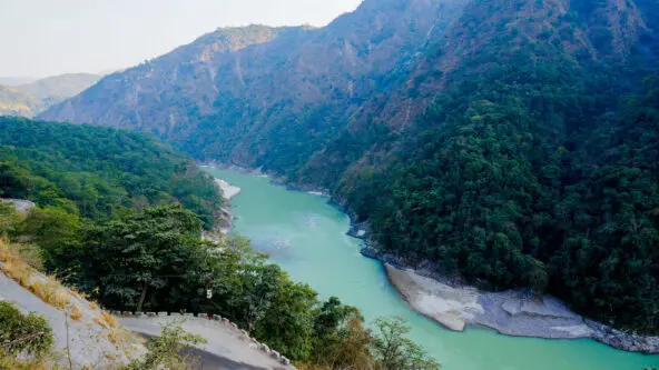 A winding river with turquoise water flows between lush green hills and steep mountains, surrounded by dense forest, under a clear sky.