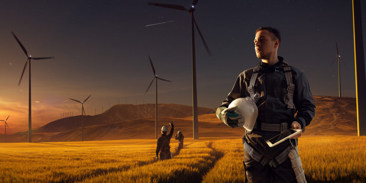 A worker in a safety harness holds a helmet and tablet, standing in a golden field with wind turbines at sunset. Two other workers consult in the background; rolling hills and a starry sky are visible.