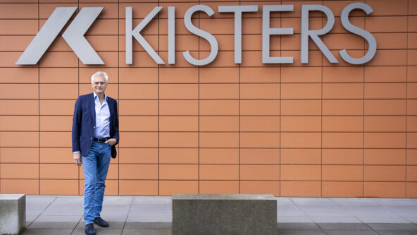 A man in a blue blazer and jeans stands in front of an orange tiled wall with a large KISTERS sign and logo. Concrete benches are on the pavement beside him.