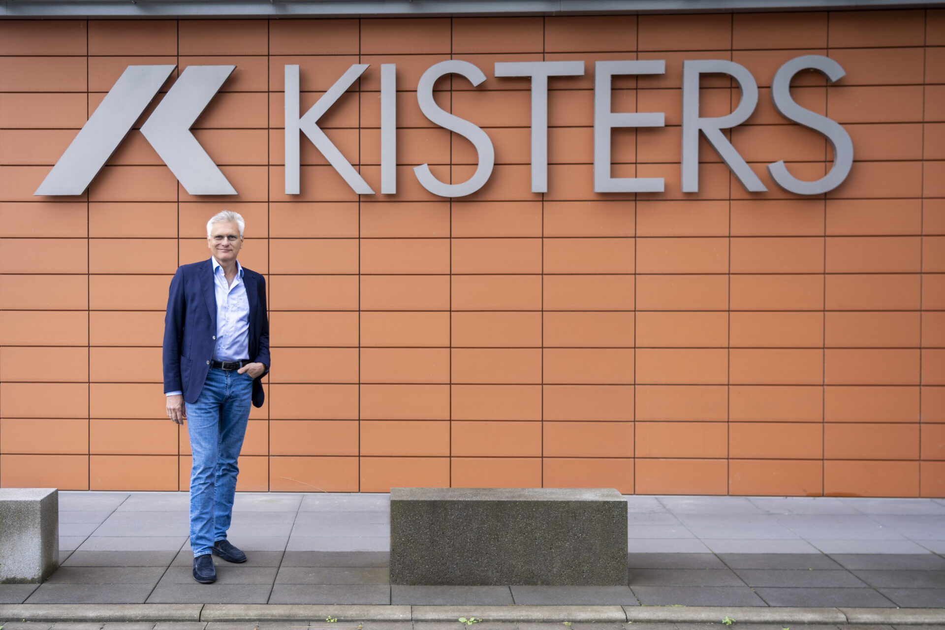 A man in a blue blazer and jeans stands in front of an orange tiled wall with a large KISTERS sign and logo. Concrete benches are on the pavement beside him.
