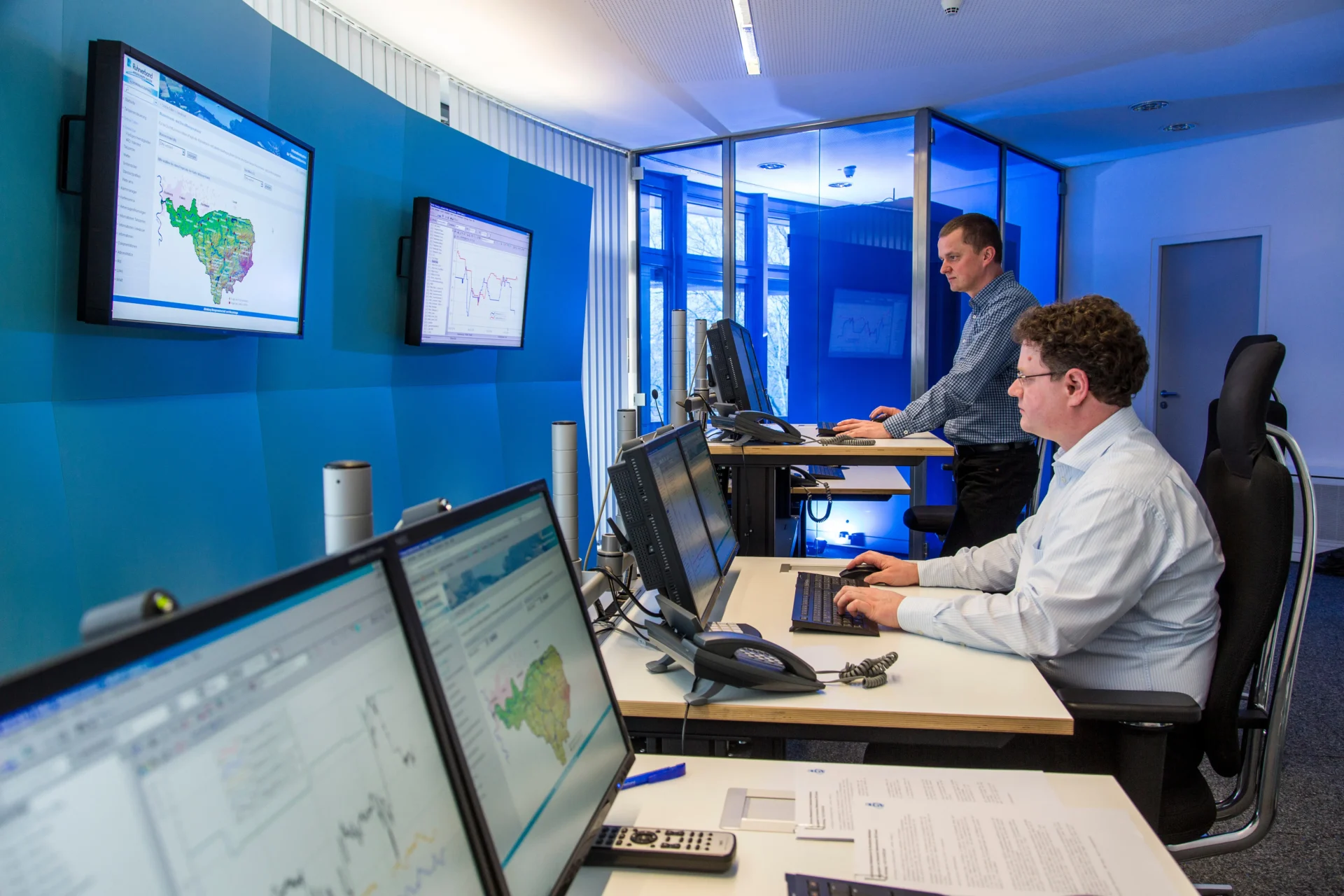 Two men work at computers in a modern office with multiple monitors displaying maps, charts, and data. One man is seated, and the other is standing at a height-adjustable desk. The room has blue walls and glass partitions.