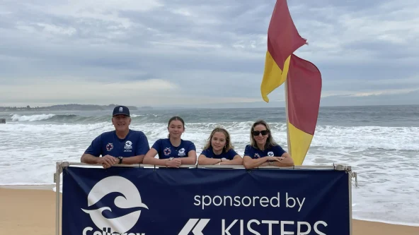 Four people in blue shirts stand behind a Collaroy Breakers sponsored by Kisters sign on a beach near a yellow-red flag. The ocean and cloudy sky are in the background.