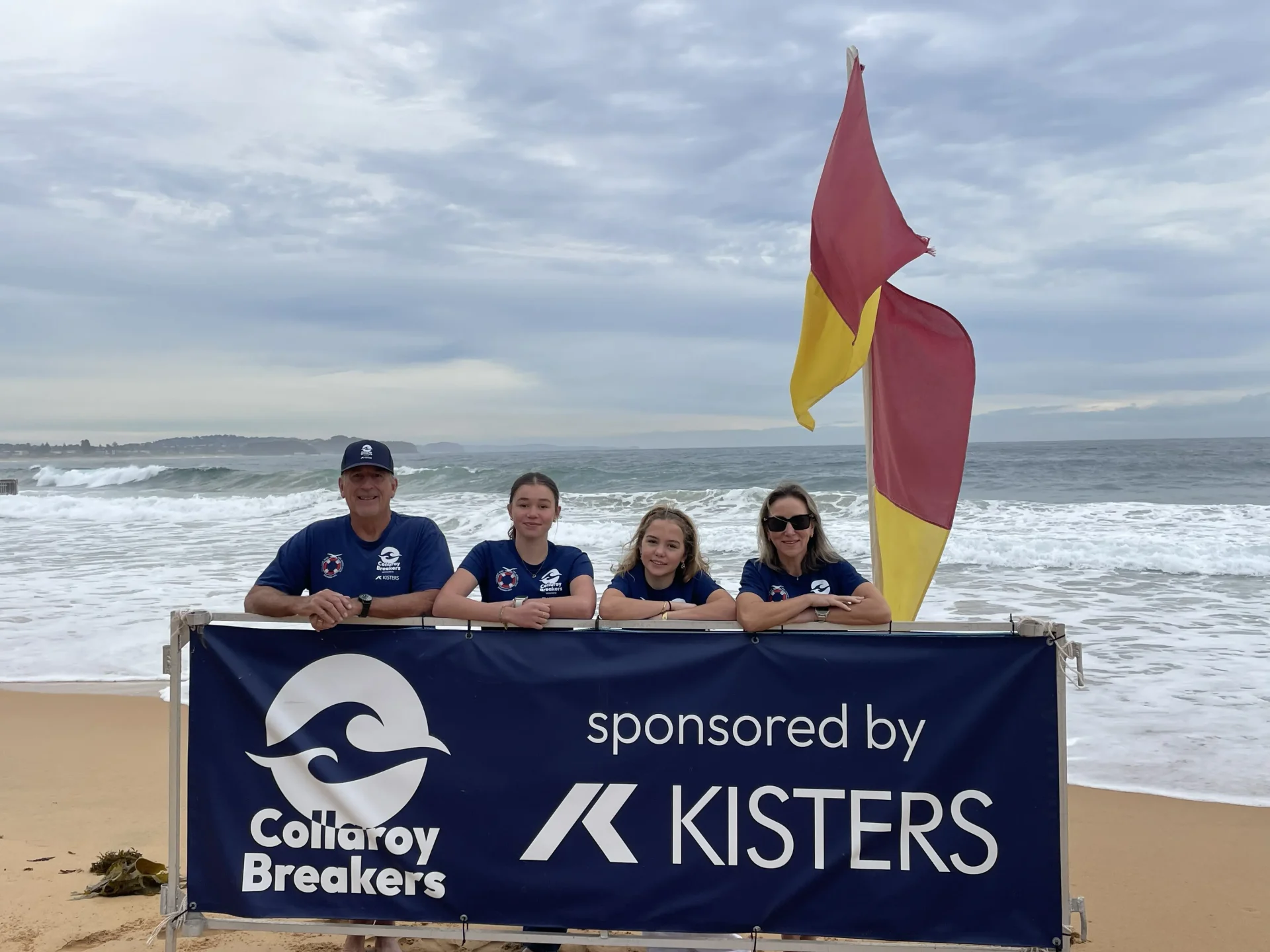 Four people in blue shirts stand behind a Collaroy Breakers sponsored by Kisters sign on a beach near a yellow-red flag. The ocean and cloudy sky are in the background.