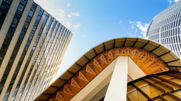 View of modern buildings from below, featuring a curved structure with wooden and glass panels, against a blue sky with scattered clouds. Two tall buildings frame the scene on each side.